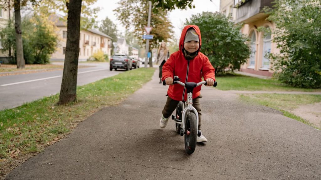 Child in a red jacket riding a balance bike on a tree-lined urban sidewalk. Autumn setting.